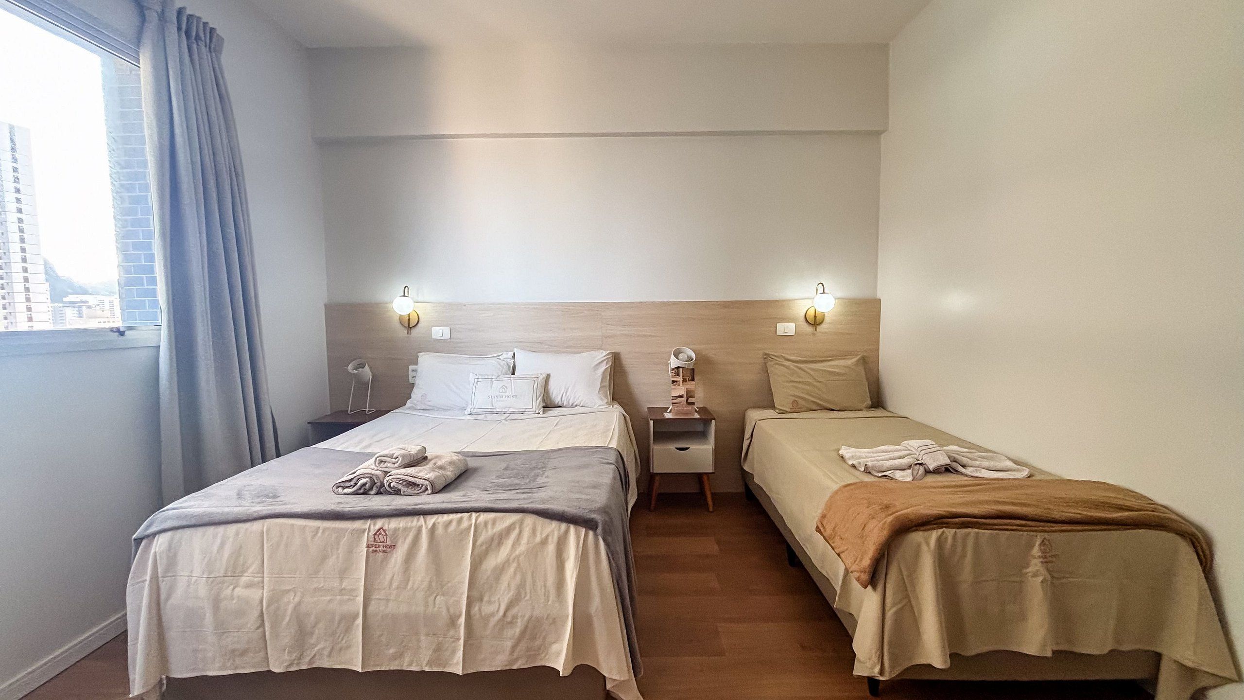 Habitación de hotel con dos camas, una mesita de noche, una lámpara y un ventanal con cortinas.