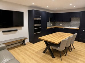 A modern kitchen with dark cabinetry, a wooden dining table, and a bench. - Stylish 1 Bedroom Town Centre Flat in Saltash 5 Mins from Train Station (Saltash)