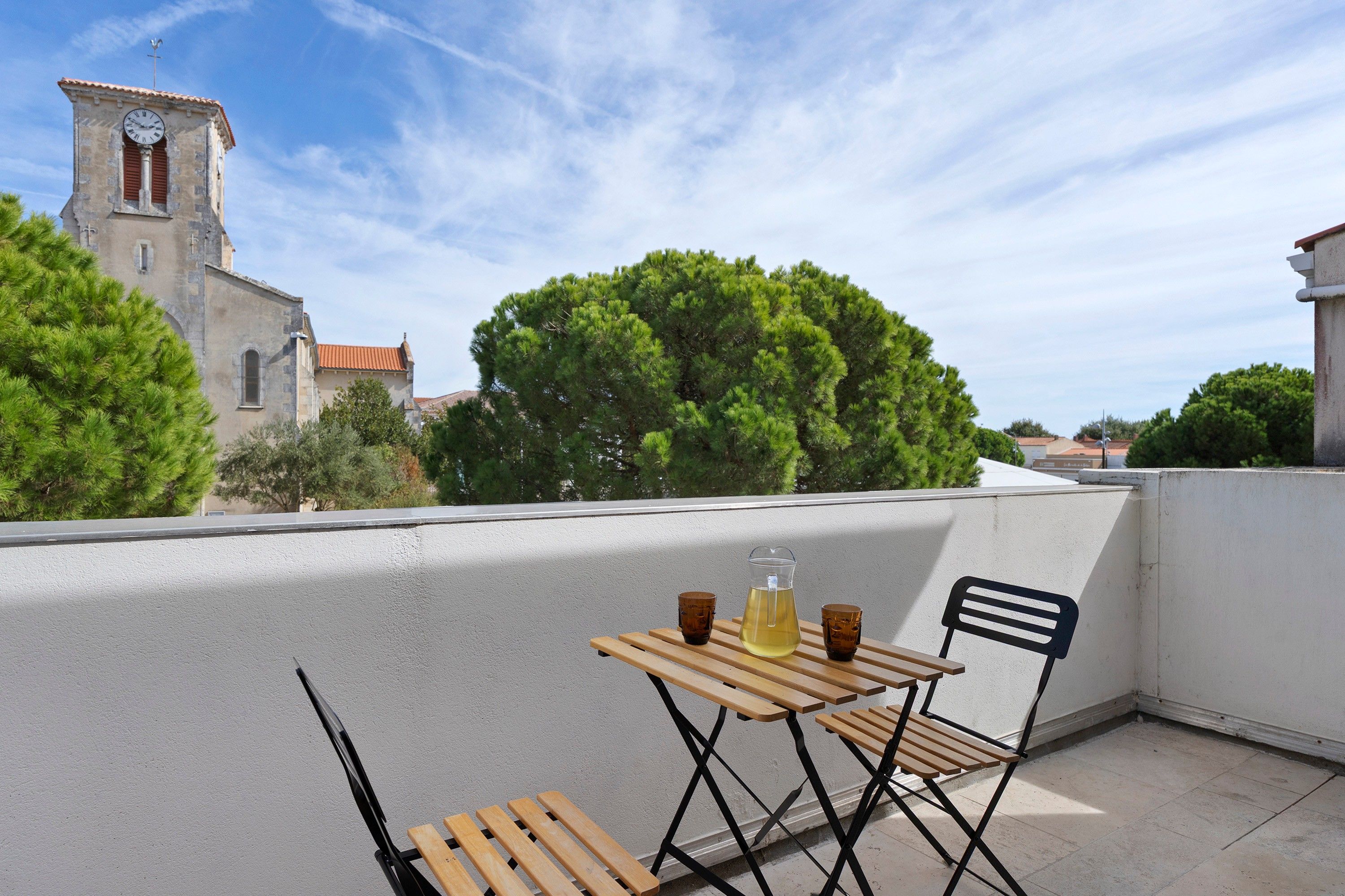 Een dakterras met een tafel en stoelen, een kan met drank, en uitzicht op een kerk en bomen.