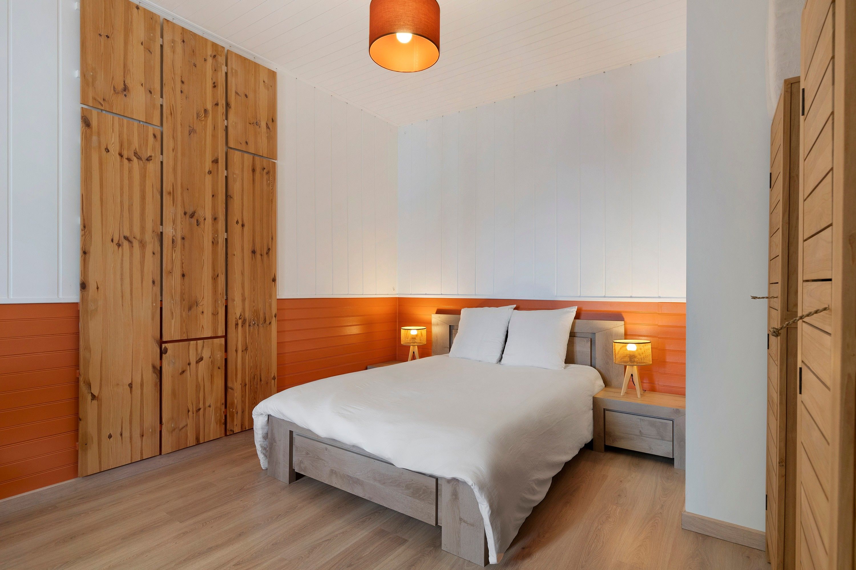 A bedroom with a wooden wardrobe, a bed with white bedding, two bedside lamps, and a wooden floor.