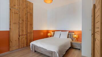 A bedroom with a wooden wardrobe, a bed with white bedding, two bedside lamps, and a wooden floor.