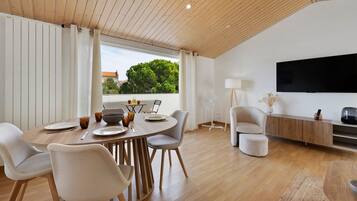 A modern living room with a dining area, wooden flooring, a flat-screen TV, and a view of greenery outside.