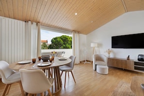 Un salon moderne comprenant un coin repas, un plancher en bois, un téléviseur à écran plat et une vue sur la verdure environnante.