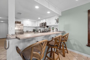 A kitchen with white cabinets, a microwave, a coffee maker, and a bar area with wicker chairs. - Castaways 4A (Gulf Shores)