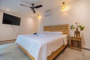 Una habitación de hotel con una cama, un ventilador de techo, un televisor de pantalla plana y una mesita de noche con una planta.