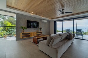 A modern living room with a herringbone-patterned wall, a flat-screen TV mounted above a wooden console, a ceiling fan, and a large window offering a view of the outdoors. - Modern family friendly, ocean view house near Dominical - Wi-Fi, inifinity pool (Dominical)