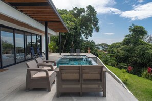 Zona de piscina moderna al aire libre con mobiliario de mimbre y vistas al follaje circundante.
