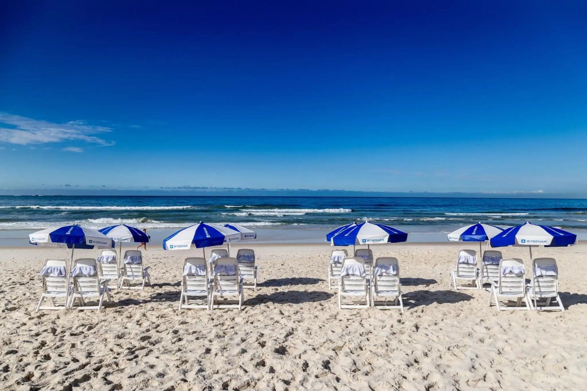 Uma praia com fileiras de cadeiras de praia brancas e guarda-sóis azuis e brancos, alguns com a marca 'WYNDHAM'.