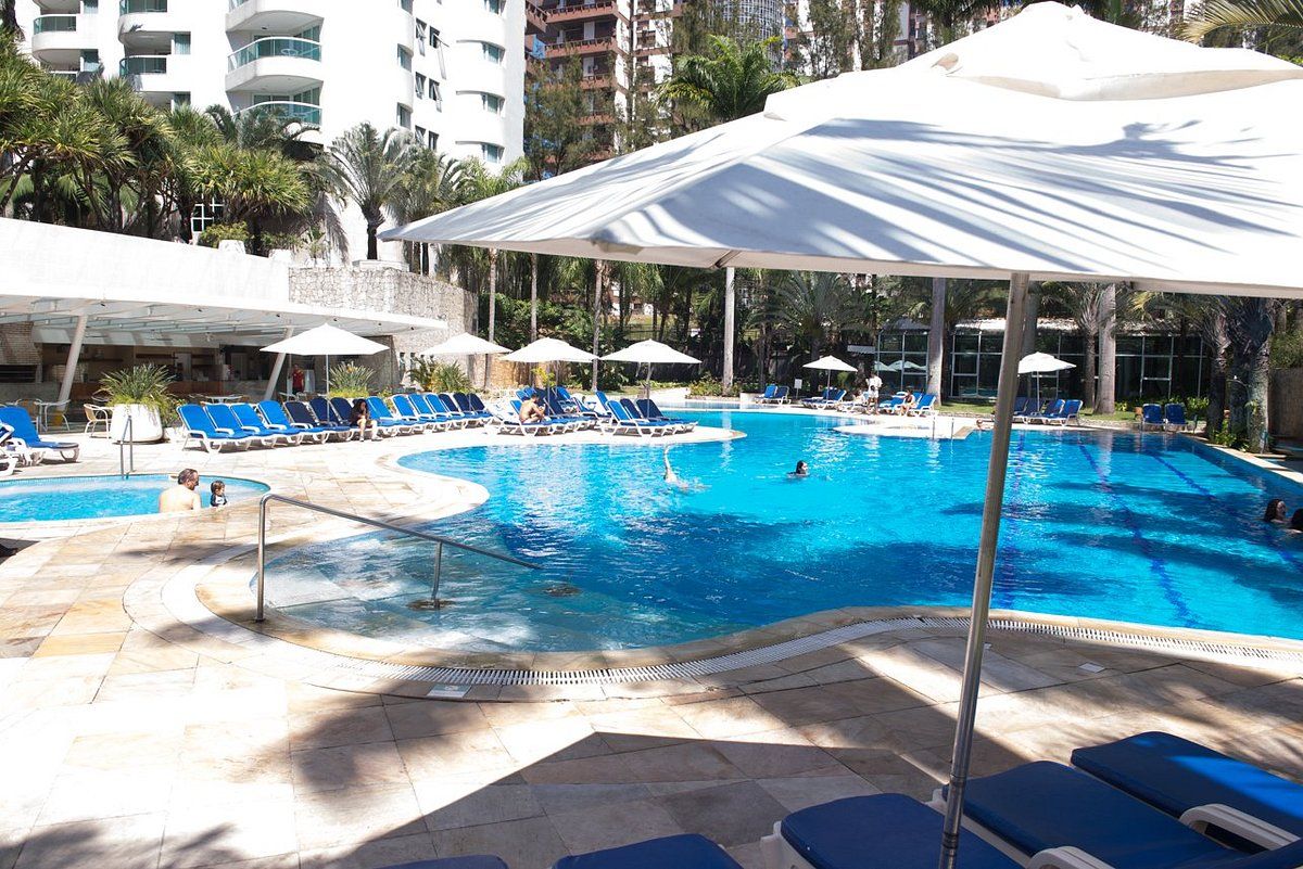 A swimming pool with lounge chairs and umbrellas, surrounded by palm trees and a building in the background.