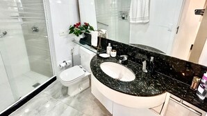 A modern bathroom with a glass shower enclosure, a white toilet, a black marble sink with a large mirror, and a potted plant. - Flat Beira Mar Barra da Tijuca Rio  (Rio de Janeiro)