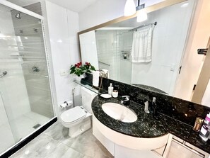 Un baño moderno con cabina de ducha de vidrio, inodoro blanco, lavabo de mármol negro y una planta en maceta.