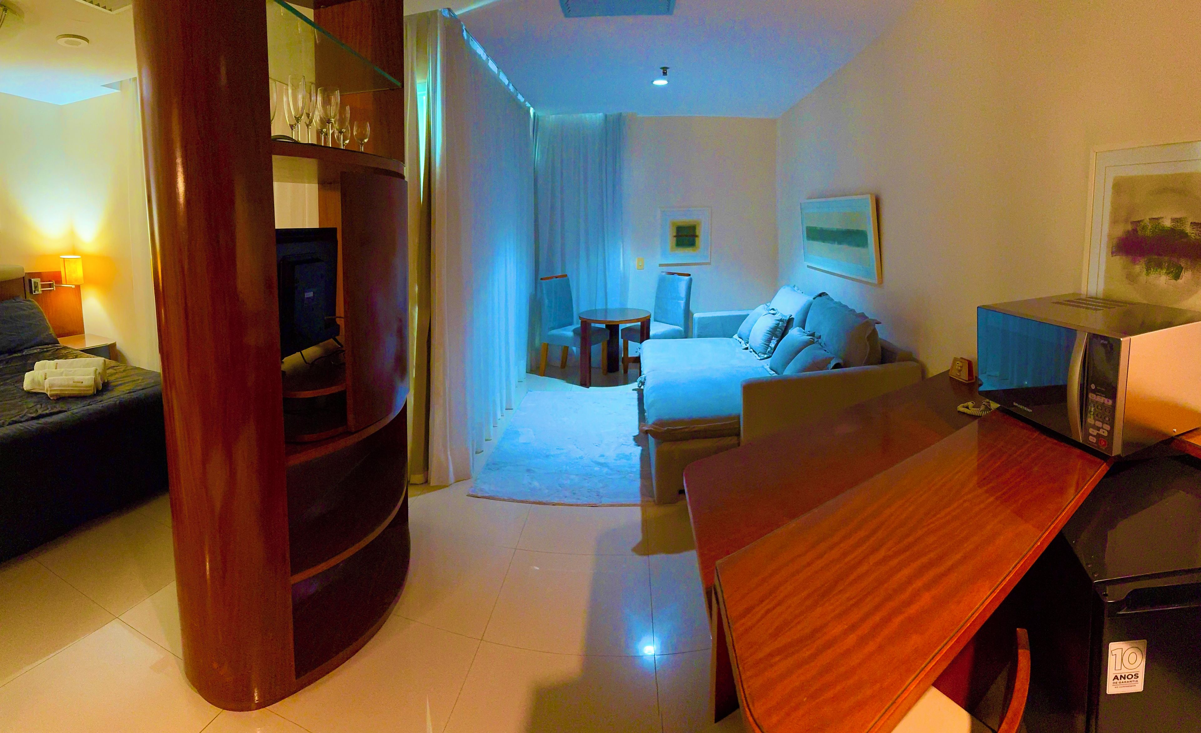 Quarto de hotel com cama, escrivaninha, cadeira, televisão e minibar.