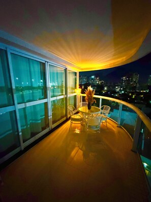A balcony with a glass railing, a round table with chairs, and a view of a cityscape at night.