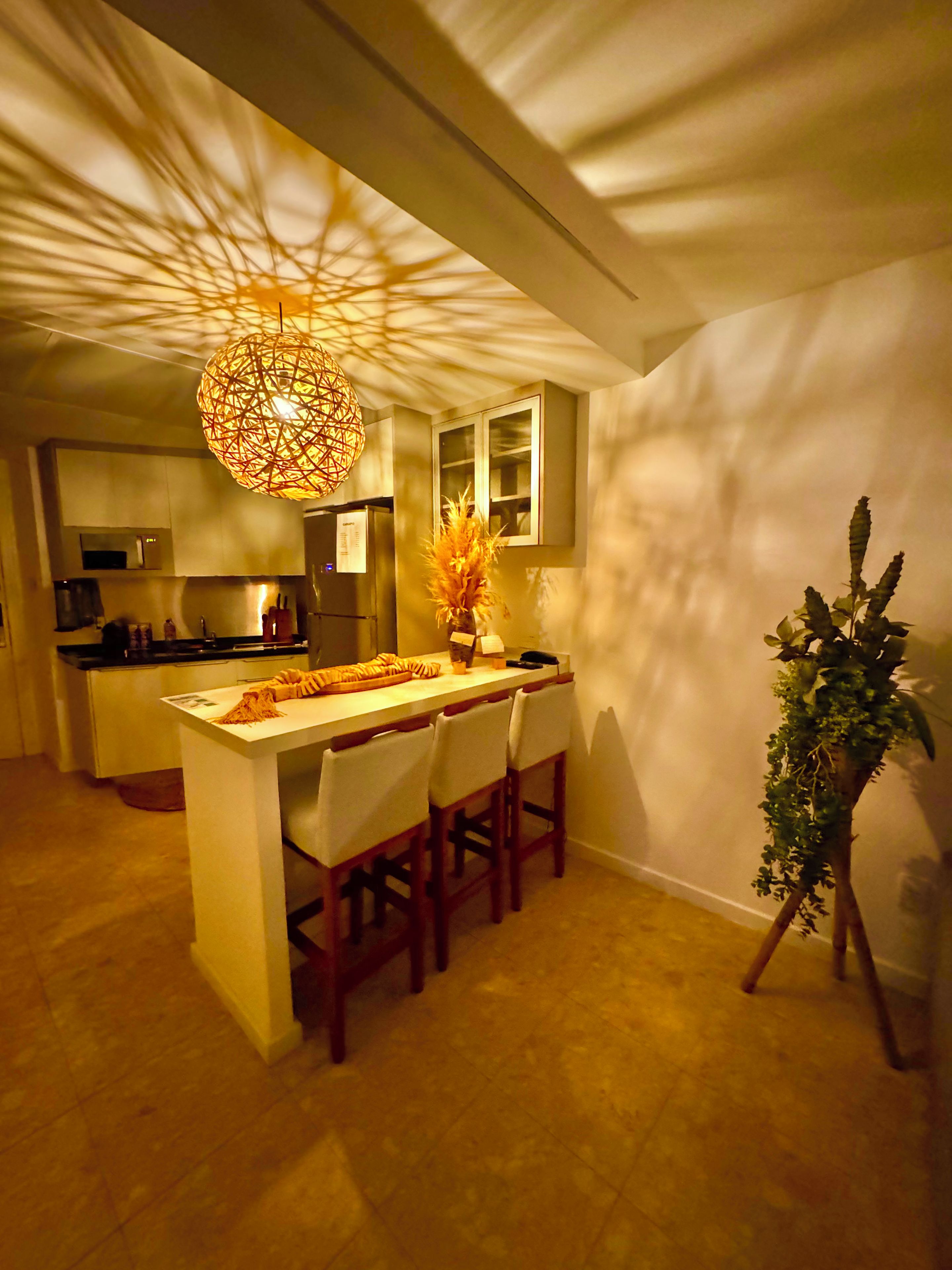 A modern kitchen with a central island, bar stools, a hanging light fixture, and a potted plant.