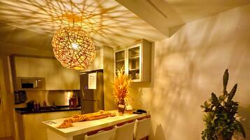 A modern kitchen with a central island, bar stools, a hanging light fixture, and a potted plant.