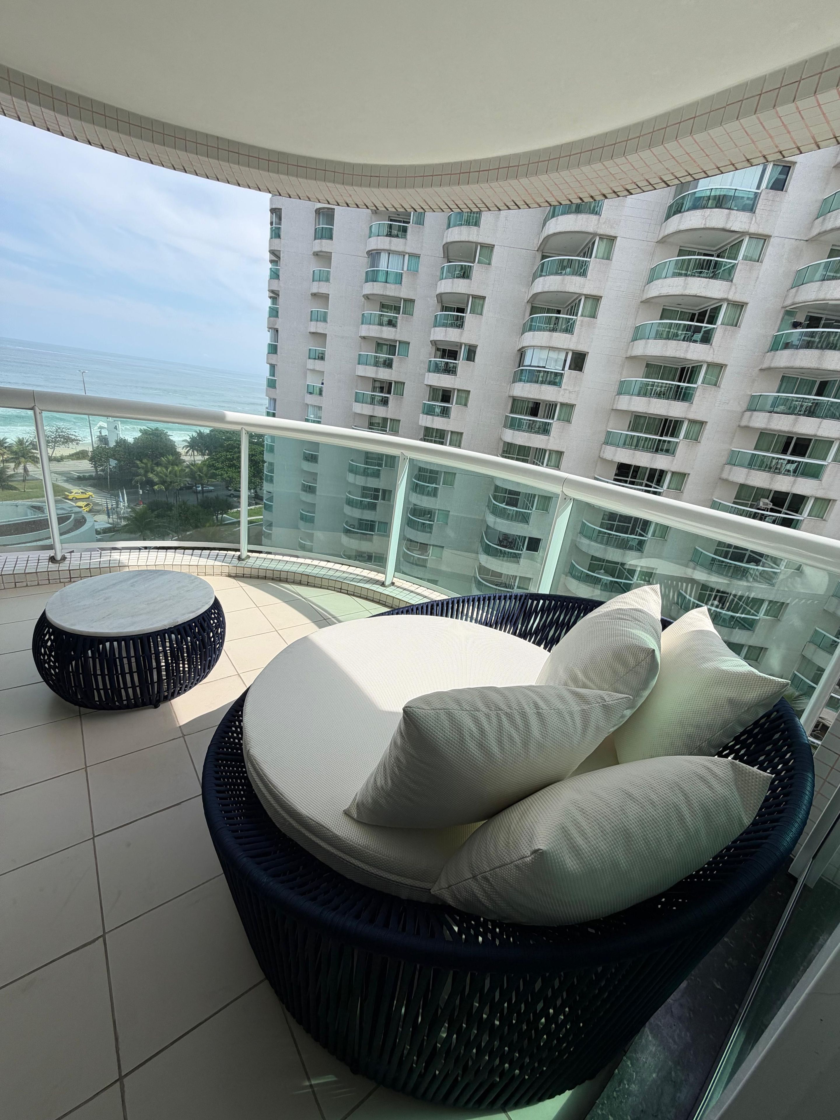A balcony with a round wicker sofa and a small round table, overlooking a high-rise building and a beach.
