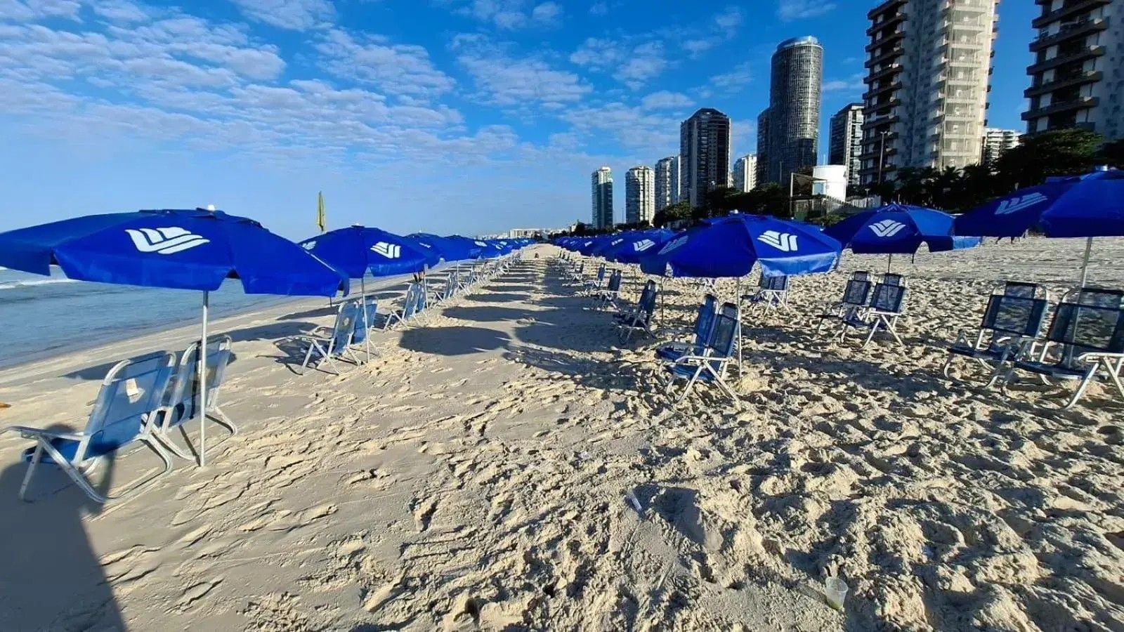 A beach with rows of blue umbrellas and chairs, a sandy shore, and tall buildings in the background.