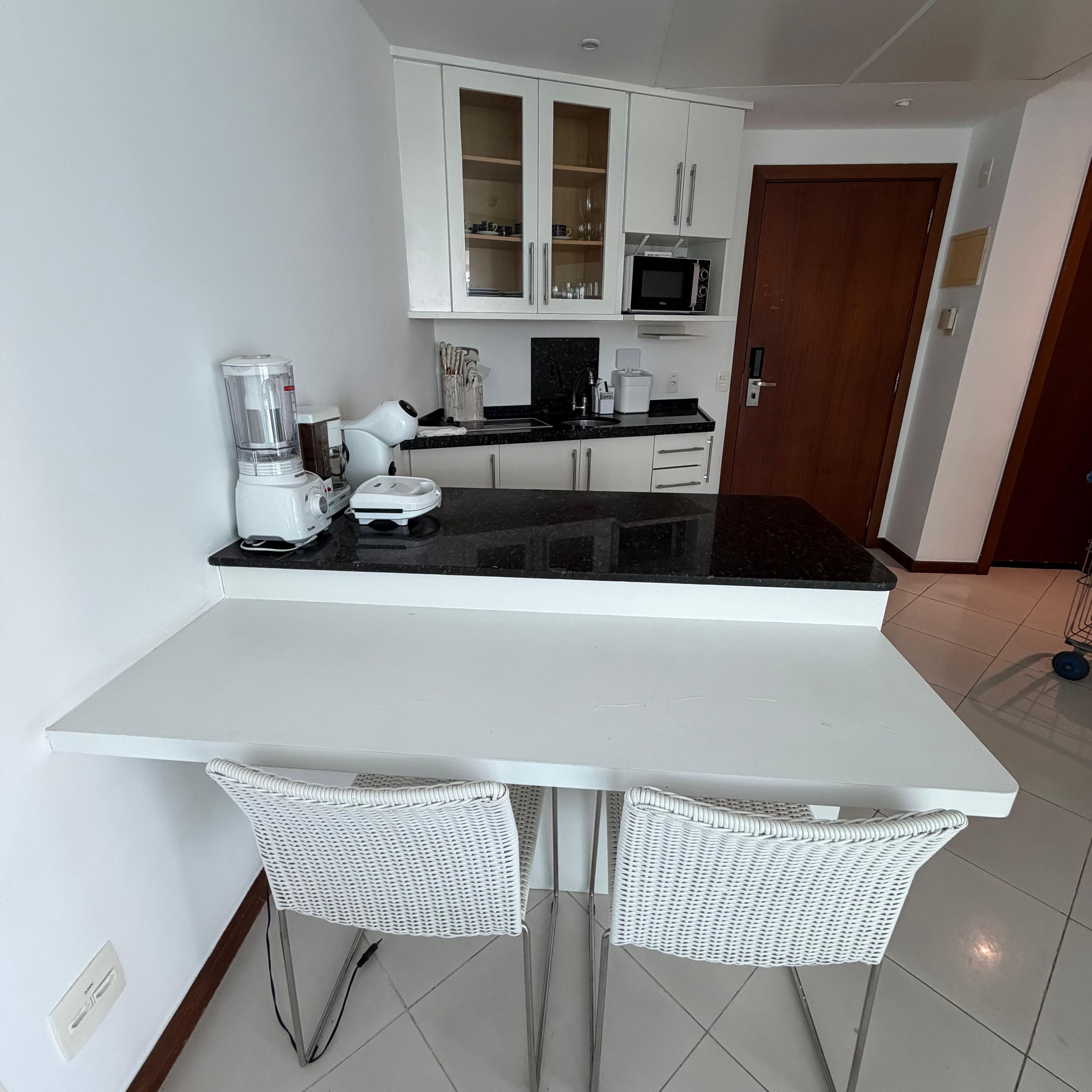 Cozinha moderna com armários brancos, bancada preta e duas cadeiras de vime.