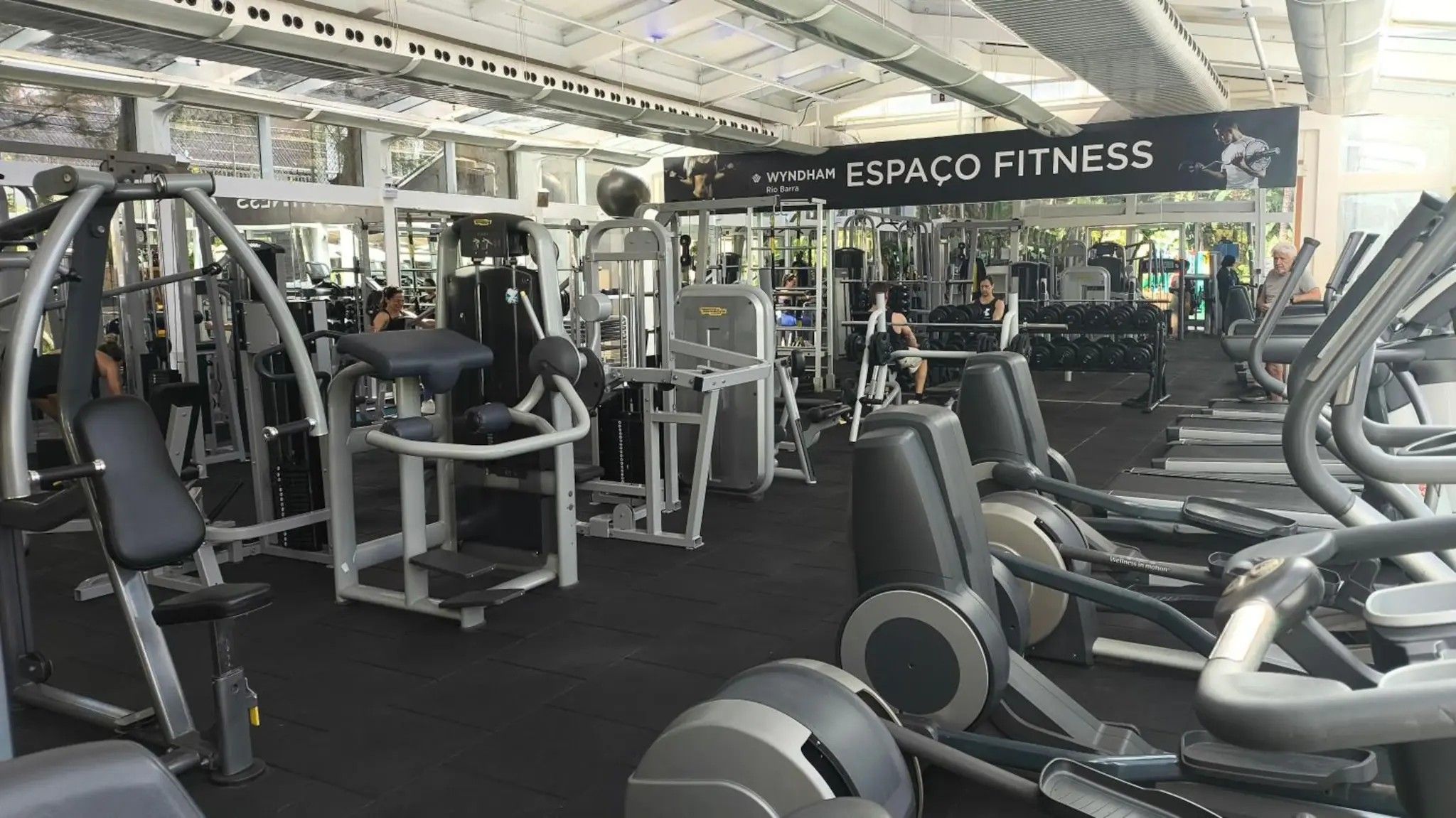 A gym with various exercise equipment, a banner reading 'WYNDHAM ESPAÇO FITNESS,' and a large window allowing natural light.