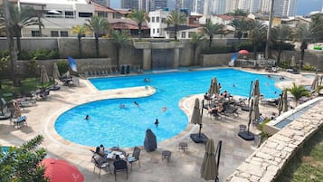 Una amplia piscina exterior con varias pistas de natación, rodeada de tumbonas y sombrillas, con edificios residenciales de fondo.