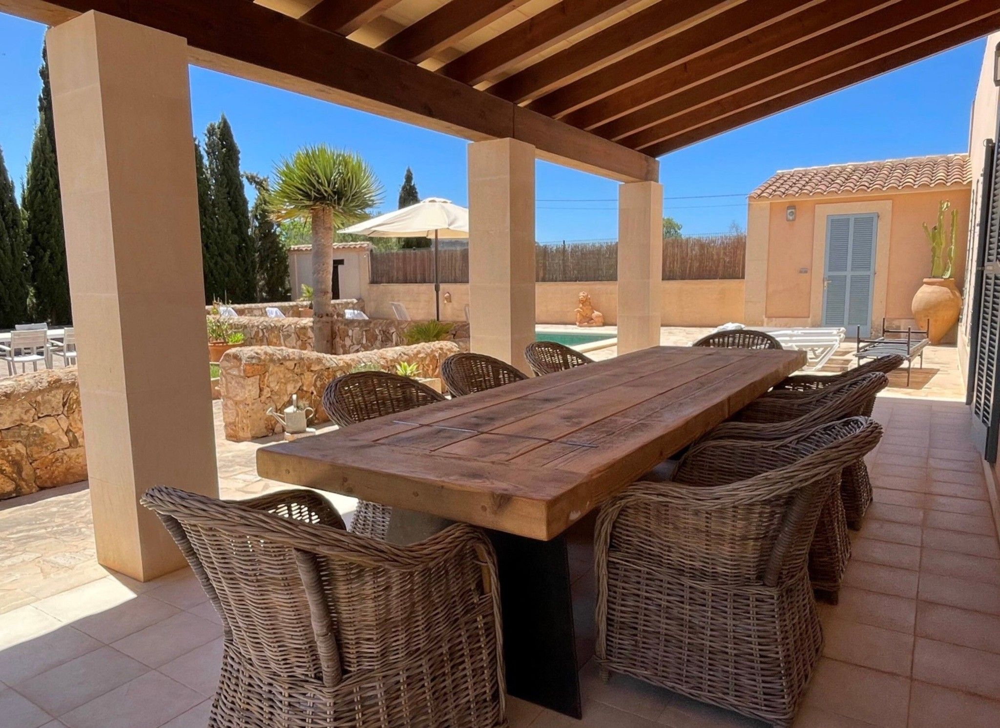 An outdoor patio with a wooden table and wicker chairs, a stone wall, and a view of a building with a tiled roof.