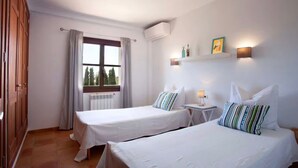 Ein Hotelzimmer mit zwei Betten, einem Fenster mit Vorhängen, einer Klimaanlage, einem Nachttisch mit Lampe und einem an der Wand befestigten Regal mit Dekoration.