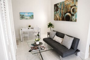 A modern living room with a grey sofa, a glass coffee table, a dining area with a white table and chairs, and a large abstract painting on the wall.
