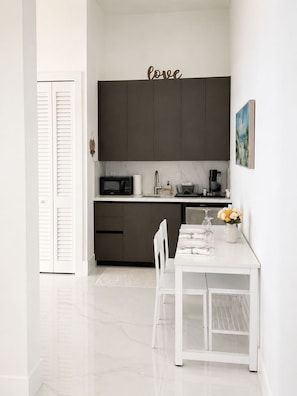 Une cuisine moderne avec des meubles de couleur sombre, un plan de travail blanc et un coin repas comprenant une table et des chaises blanches.