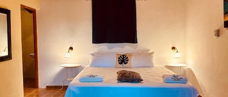 A bedroom with a bed, two bedside tables, a nightstand, a wooden ceiling, and a wall-mounted light fixture.