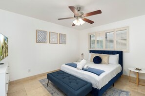 Une chambre à coucher avec un lit, un ventilateur de plafond, une fenêtre avec des stores et un téléviseur fixé au mur.