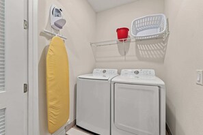 Un espace dédié à la lessive, avec deux machines à laver blanches, une planche à repasser jaune, un seau rouge et un panier à linge blanc, le tout placé sur une étagère fixée au mur.