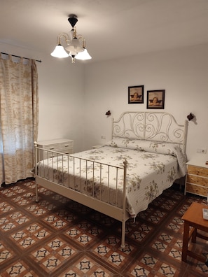 Un dormitorio con cama, mesita de noche, una mesa, cortinas estampadas y una lámpara de techo.