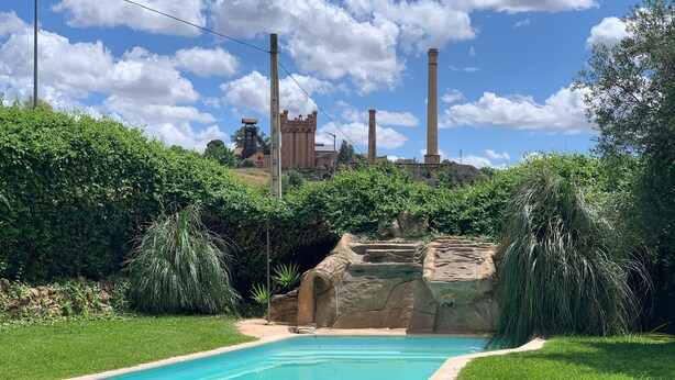 En pool omgivet af frodig vegetation, med udsigt til fjerne skorstene og en borglignende bygning.