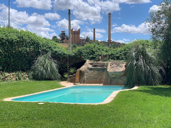 Una piscina rodeada de exuberante vegetación, con una vista distante de chimeneas industriales y una estructura que parece un castillo.