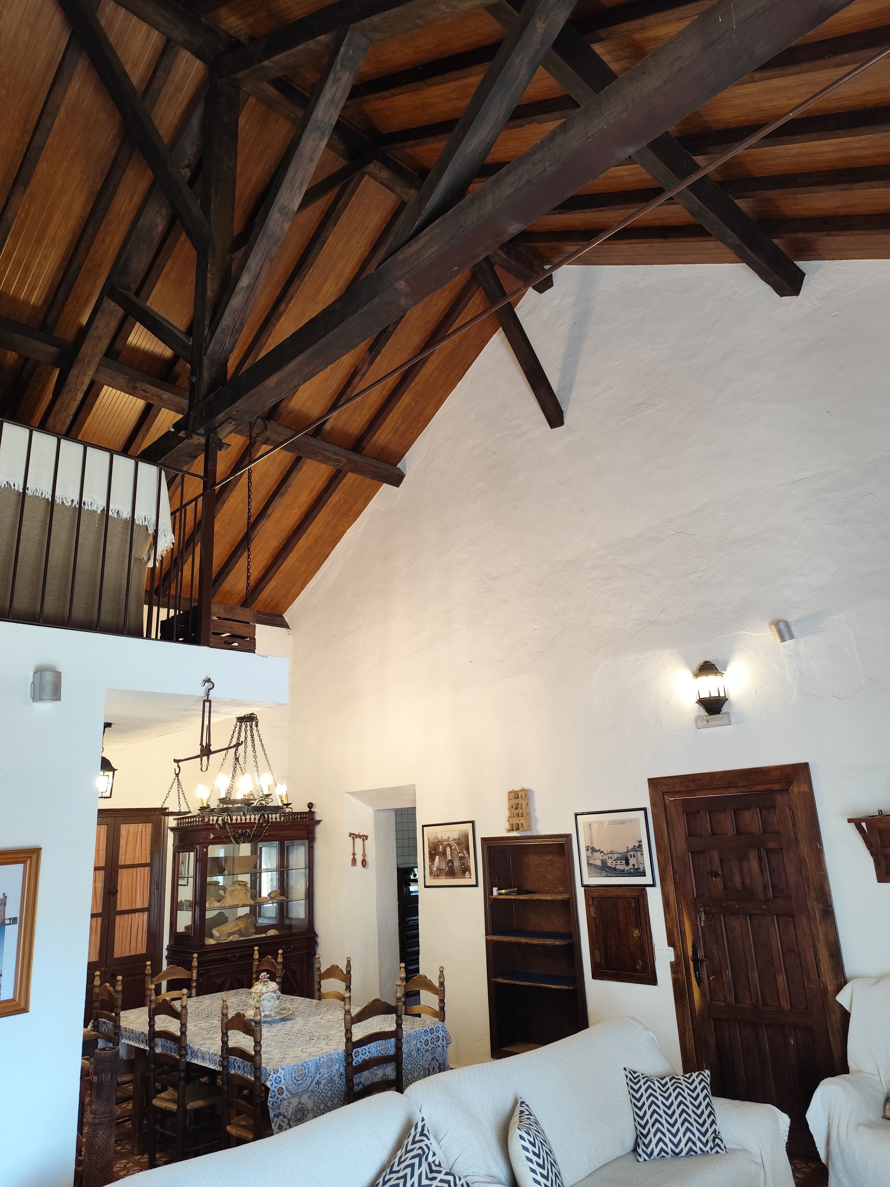 A rustic interior with exposed wooden beams, a dining area with a chandelier, and a sofa with patterned cushions.