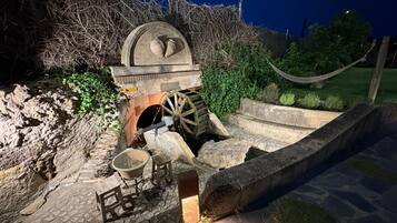 Una ruota idraulica in pietra con una panchina di legno e un secchio, circondata da mura in pietra e vegetazione.
