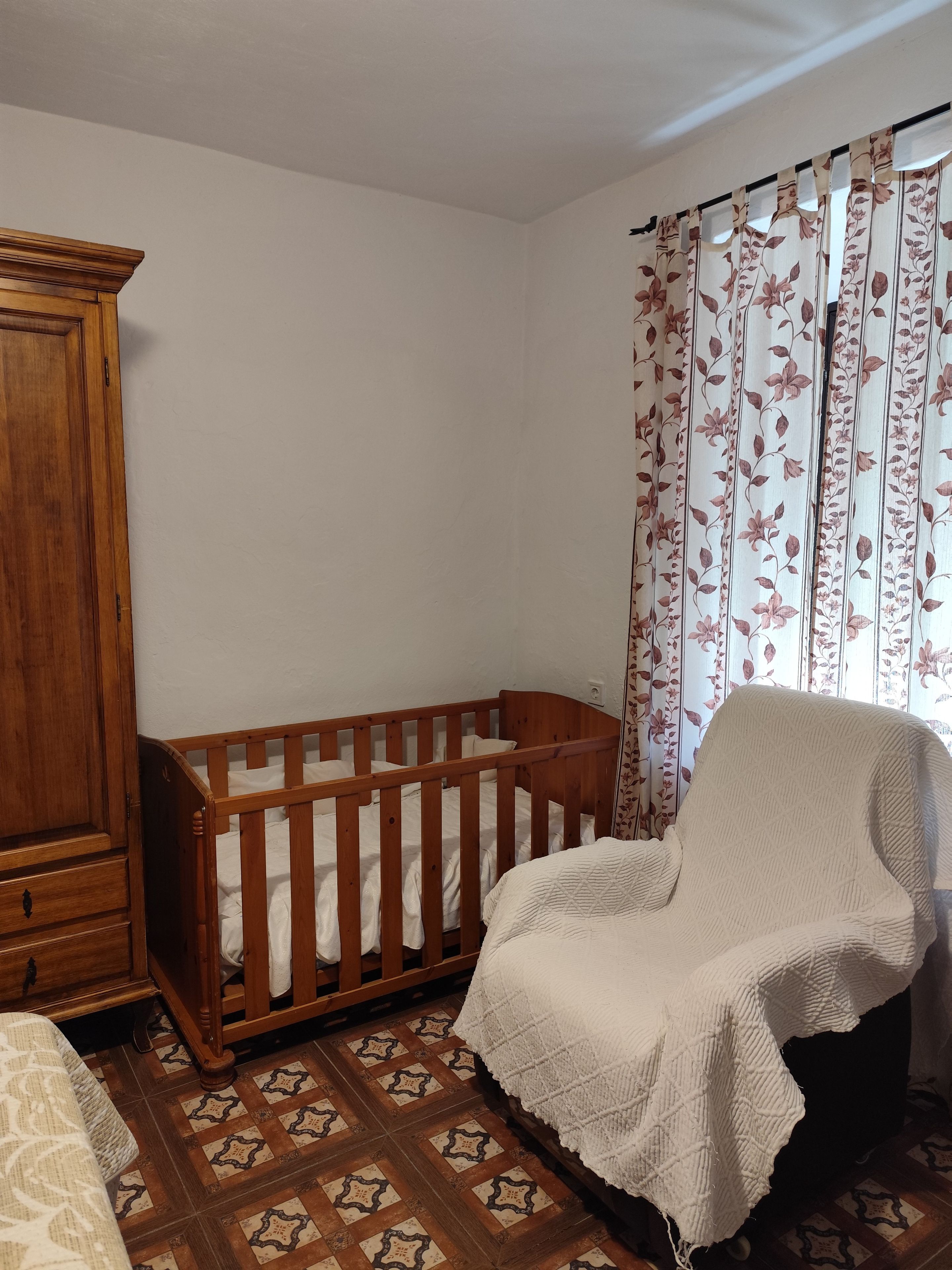 Una habitación con una cuna de madera, una silla con funda blanca y cortinas estampadas.