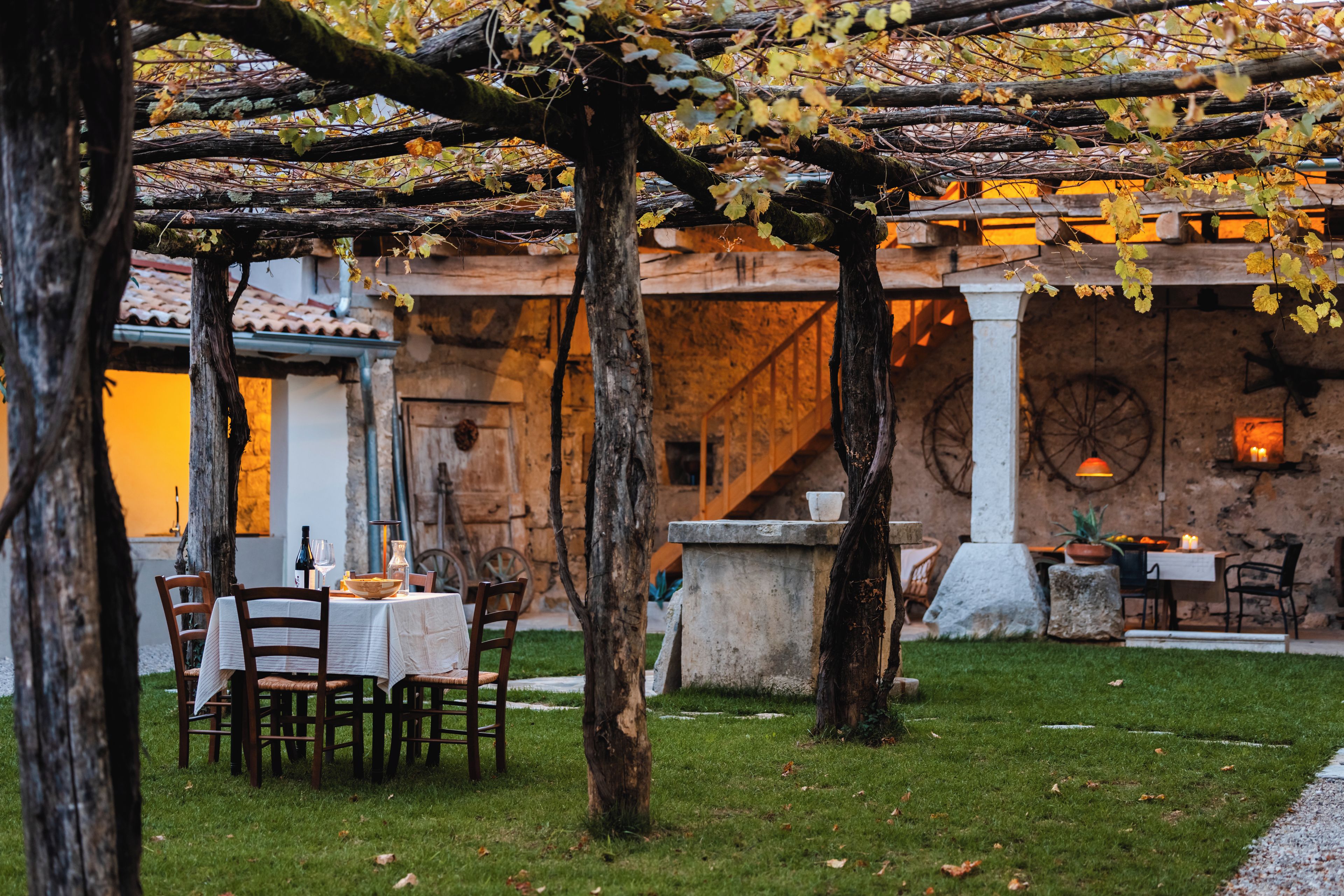 Un área de comedor al aire libre con mesas y sillas, una pérgola y un muro de piedra al fondo.