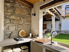 Cozinha com parede de pedra, bancada de madeira e vista para uma casa com varanda.