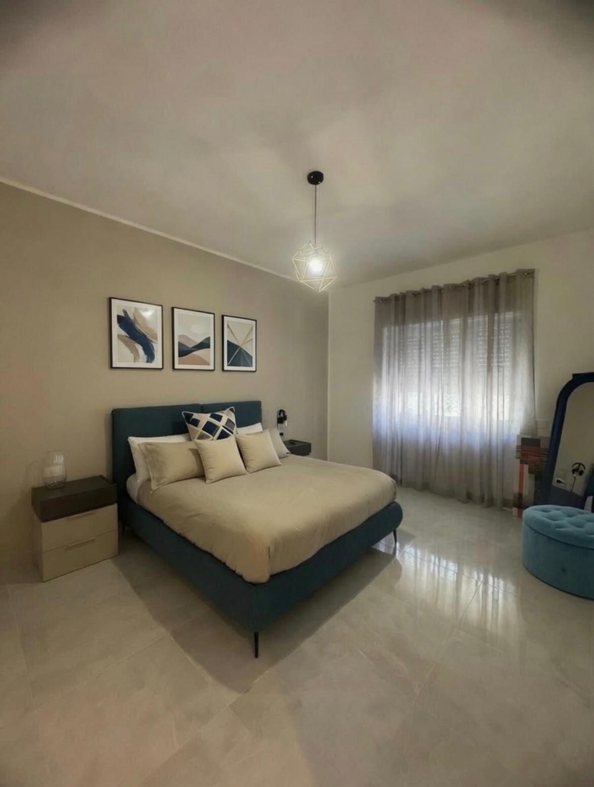 Quarto com cama, mesa de cabeceira, luminária e janela com cortinas.