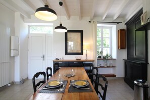 A dining area with a wooden table set for two, a mirror above the table, and a window with curtains. - Cottage 'Gite Le Sainte Marie' with Private Terrace, Private Garden and Wi-Fi (Sainte-Marie-du-Mont)
