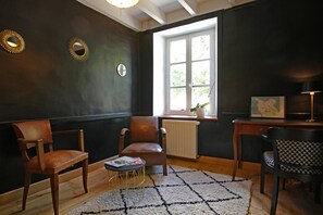 A room with a dark wall, a window, a radiator, two chairs, a small round table with a magazine, and a lamp. - Cottage 'Gite Le Sainte Marie' with Private Terrace, Private Garden and Wi-Fi (Sainte-Marie-du-Mont)