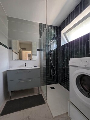 A modern bathroom with a glass shower enclosure, a built-in washing machine, and a vanity with a mirror.