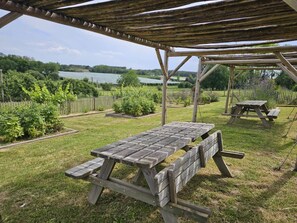 An outdoor picnic area with wooden tables and benches, surrounded by a grassy lawn and a wooden fence.