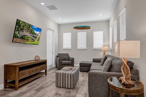 A modern living room with a flat-screen TV, a sofa, a chair, a wooden console, and a lamp. - Pickled Parrot Cottage - Private Pool Near Disney & Island H2O Access - 8024ST (Kissimmee)