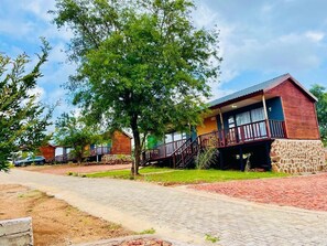 A row of wooden cabins with stone foundations, a paved walkway, and a tree in the foreground.