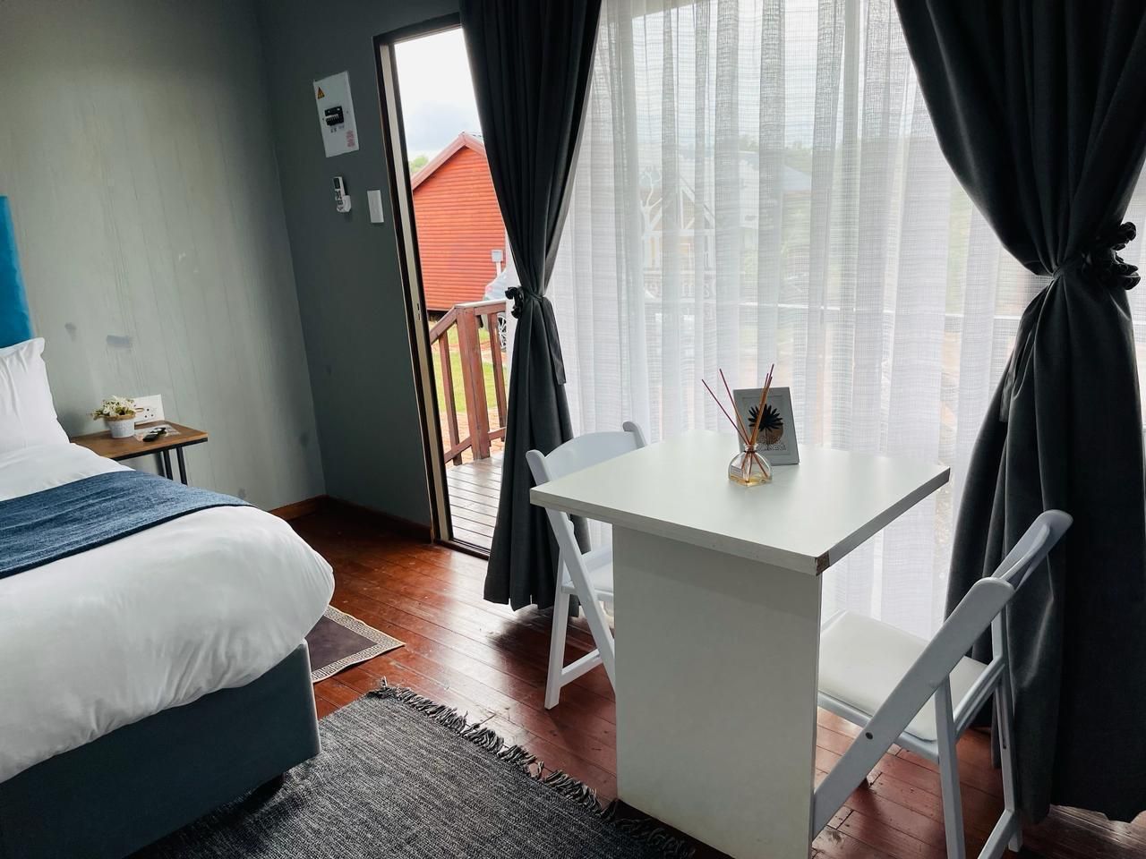 A bedroom with a bed, a small table, and a chair. There is a window with curtains, a door leading outside, and a view of a red building.