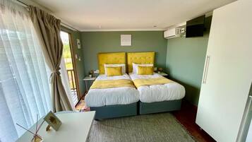 A double bed with yellow headboards and white bedding, a small table with a picture frame, a white cabinet, and a window with curtains.