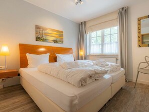 A hotel room with a large bed, a nightstand, a lamp, a window with curtains, and a painting on the wall. - "seenelke" (St. Peter-Ording)