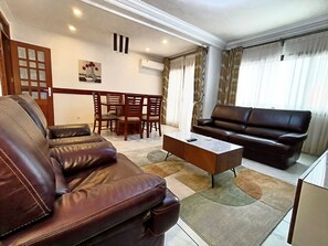 A living room with a brown leather sofa, a white coffee table, and a brown leather couch. There is a dining table with chairs in the background.